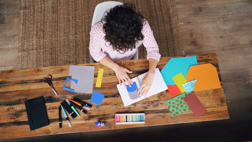 woman working on a project with a notebook, pens, scissors, and colored paper
