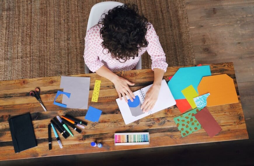 woman working on a project with a notebook, pens, scissors, and colored paper