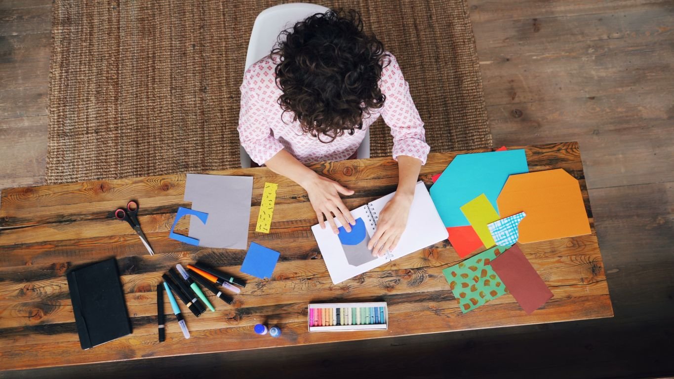 woman working on a project with a notebook, pens, scissors, and colored paper